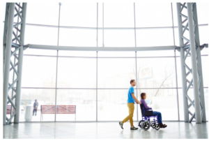 Imagen del interior de un aeropuerto con fondo acristalado en el que se ve a dos personas, una en silla de ruedas siendo empujada por otra persona andando