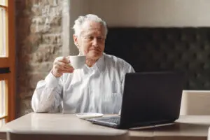 Hombre mayor tomando cafe mientras realiza tarea con ordenador portatil