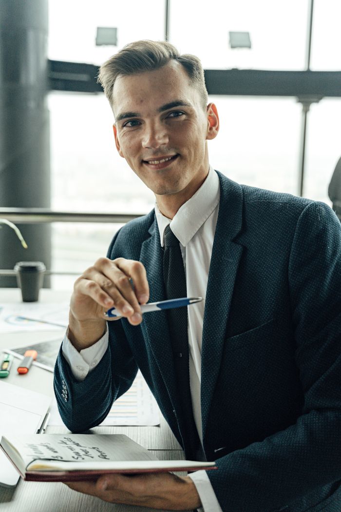 A smiling young man in a suit holds a pen and notebook in a modern office environment.