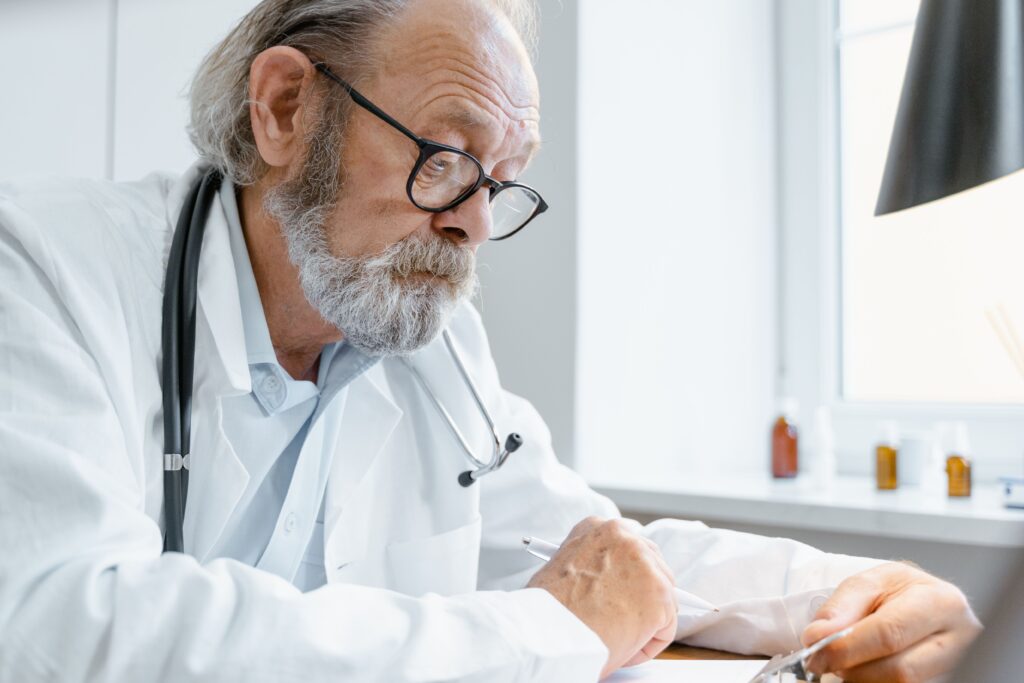 Elderly doctor wearing eyeglasses and stethoscope writes at office desk.