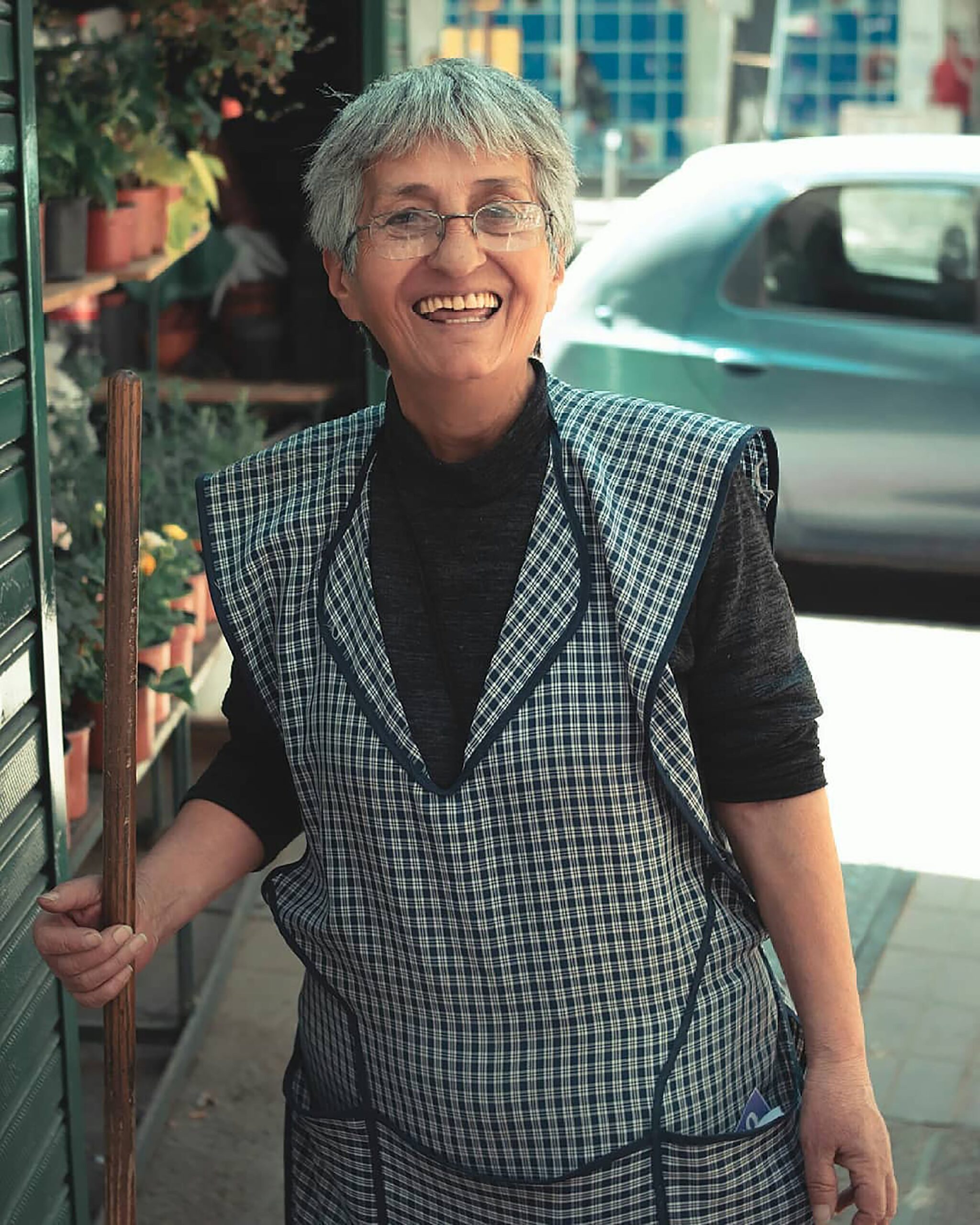 Cheerful elderly woman with eyeglasses holding a broom, outdoors in Mendoza, Argentina.